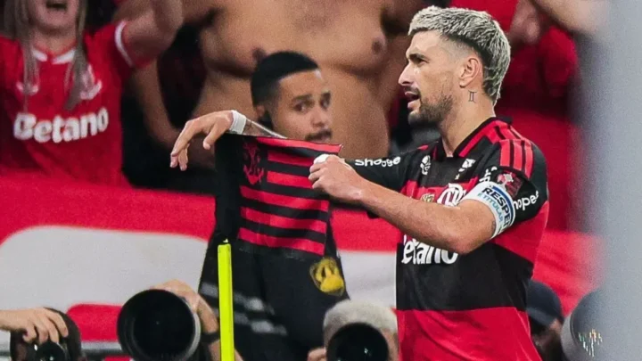 Jogador do Flamengo segura camisa da equipe durante partida, cercado por companheiros e torcedores em ambiente de estádio