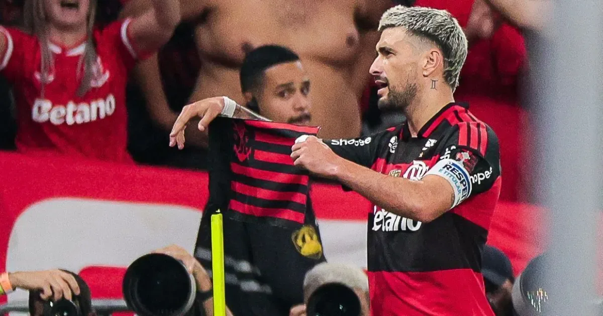 Jogador do Flamengo segura camisa da equipe durante partida, cercado por companheiros e torcedores em ambiente de estádio