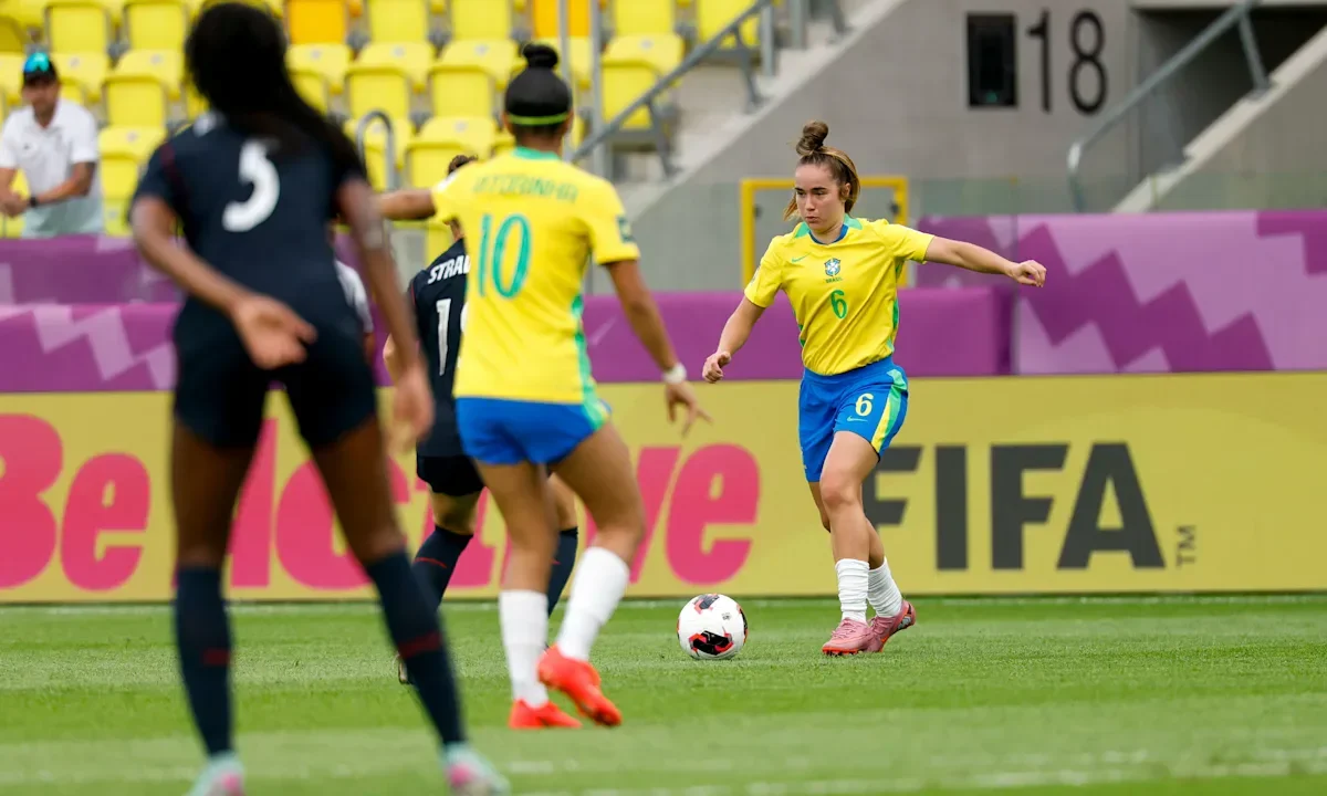 Jogadora da seleção brasileira em uniforme amarelo e azul domina a bola durante partida de futebol feminino em estádio com assentos amarelos e logo da FIFA visível