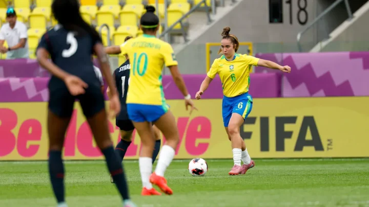 Jogadora da seleção brasileira em uniforme amarelo e azul domina a bola durante partida de futebol feminino em estádio com assentos amarelos e logo da FIFA visível