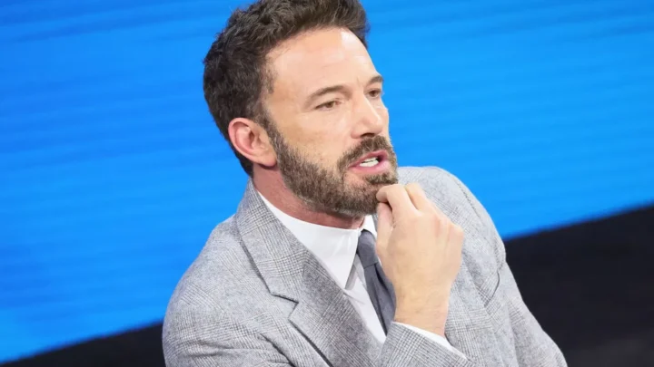 Ben Affleck, de terno cinza e barba, fala diante de fundo azul, com a mão no rosto em gesto contemplativo