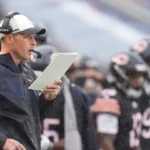 Treinador com boné e fone de ouvido segura tablet enquanto comanda estratégias em jogo de futebol americano sob chuva
