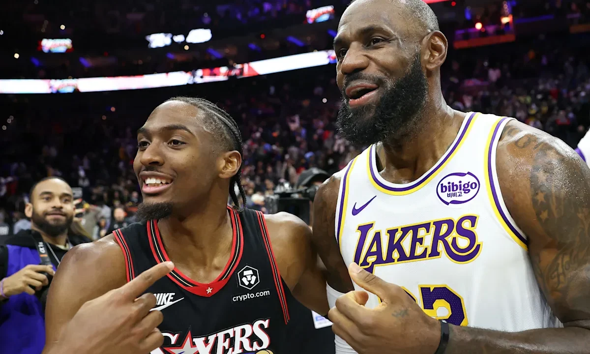 Dois jogadores de basquete em uniforme conversam na quadra durante partida da NBA, com público ao fundo e placares iluminados no teto da arena