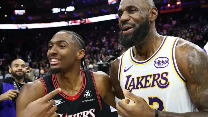 Dois jogadores de basquete em uniforme conversam na quadra durante partida da NBA, com público ao fundo e placares iluminados no teto da arena