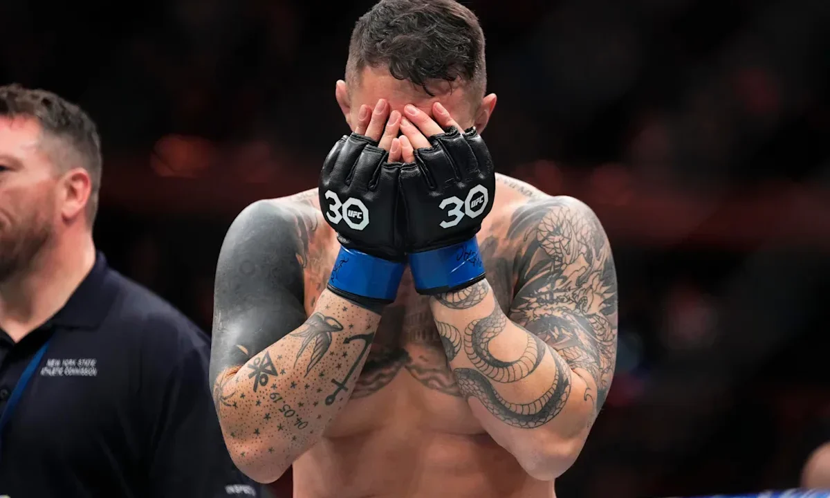 Lutador de MMA com luvas de combate preta e azul cobre o rosto com as mãos após vencer luta pelo título interim dos pesos-pesados no UFC