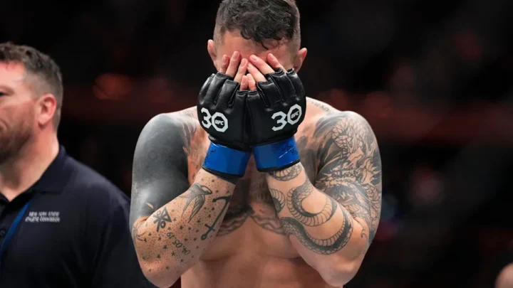Lutador de MMA com luvas de combate preta e azul cobre o rosto com as mãos após vencer luta pelo título interim dos pesos-pesados no UFC