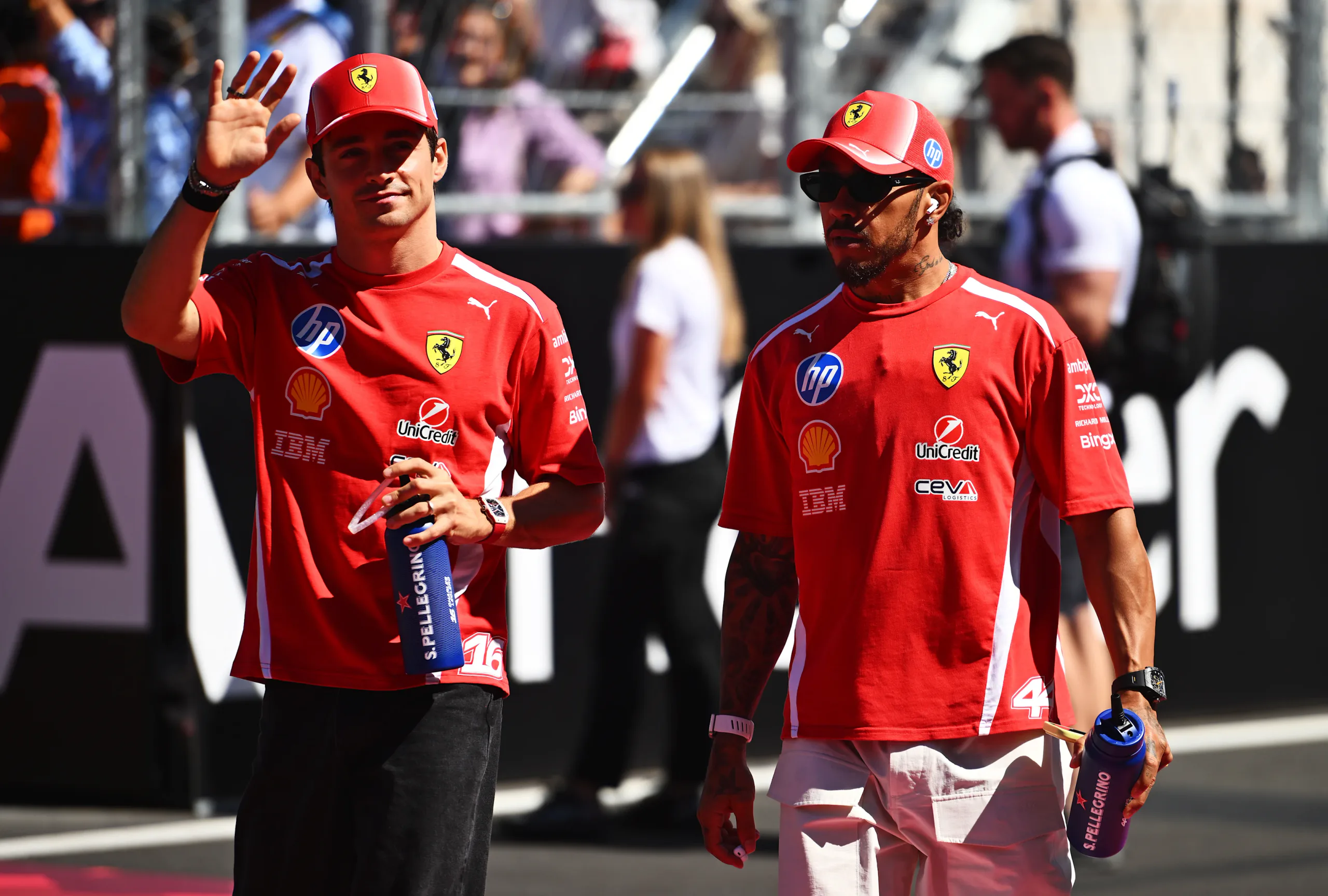 Dois pilotos de Fórmula 1 da Ferrari caminham na pista durante o paddock, com o piloto à esquerda acenando para a torcida em dia de evento da categoria