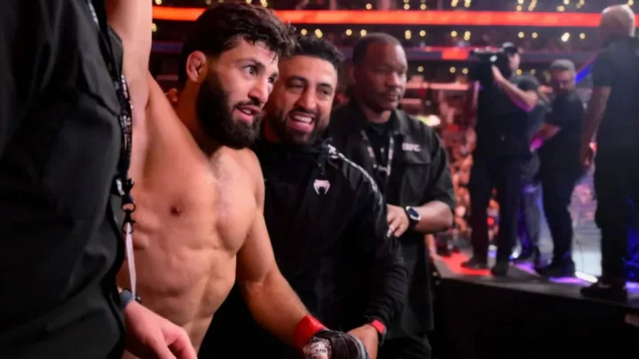 Lutador com luvas de MMA conversa com membros da equipe no octógono durante evento de UFC em arena iluminada