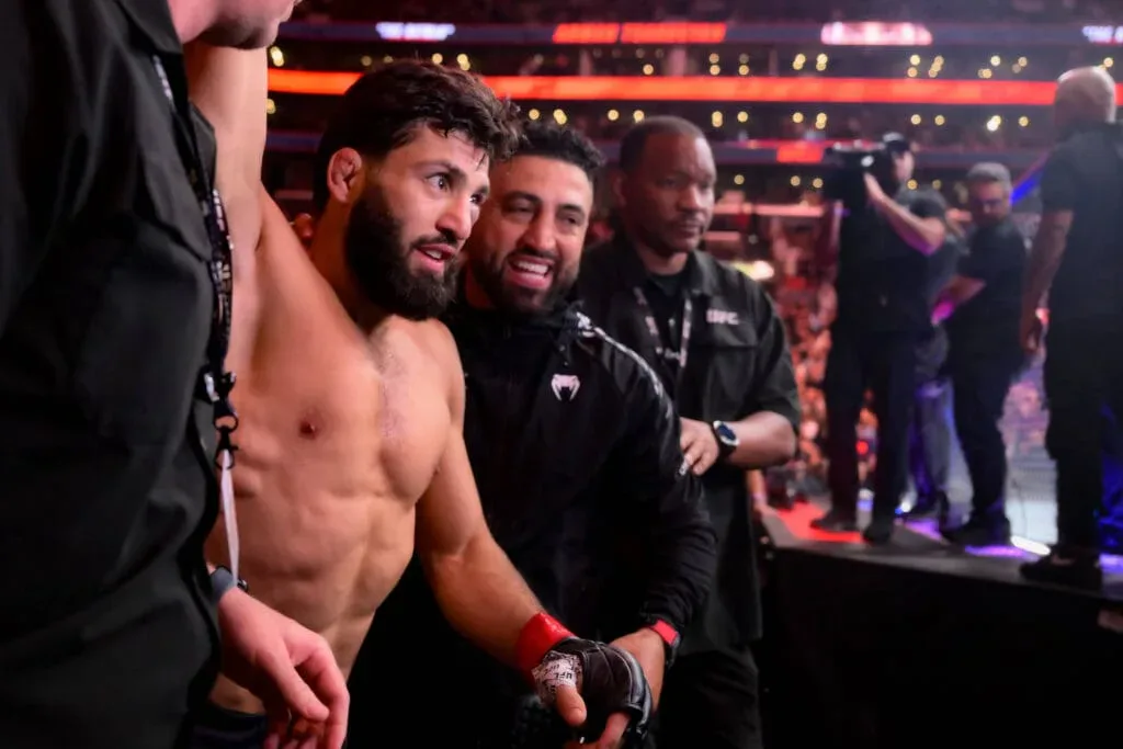 Lutador com luvas de MMA conversa com membros da equipe no octógono durante evento de UFC em arena iluminada