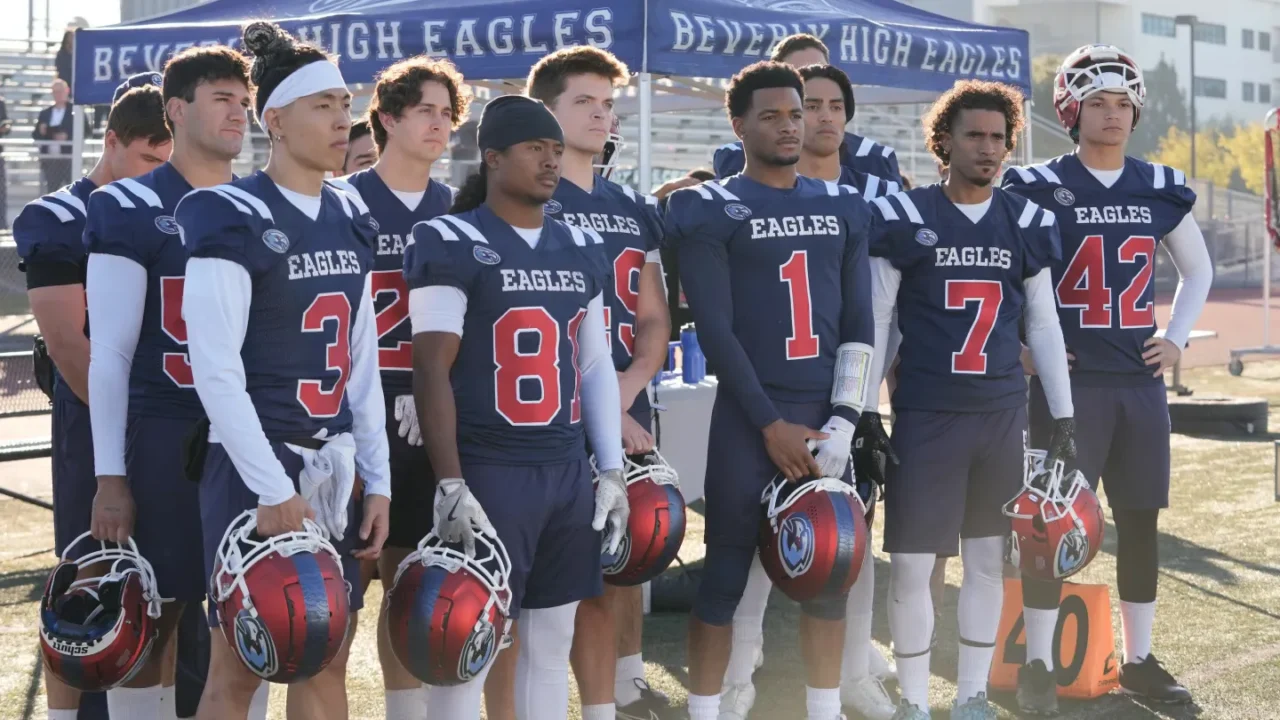 Grupo de jogadores de futebol americano da escola Beverly High Eagles, uniformizados em azul e branco com capacetes vermelhos, posam juntos no estádio antes do jogo