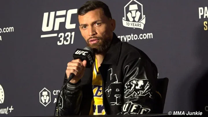 Lutador comparece a coletiva de imprensa do UFC 331 segurando microfone em frente a painel com logos do evento