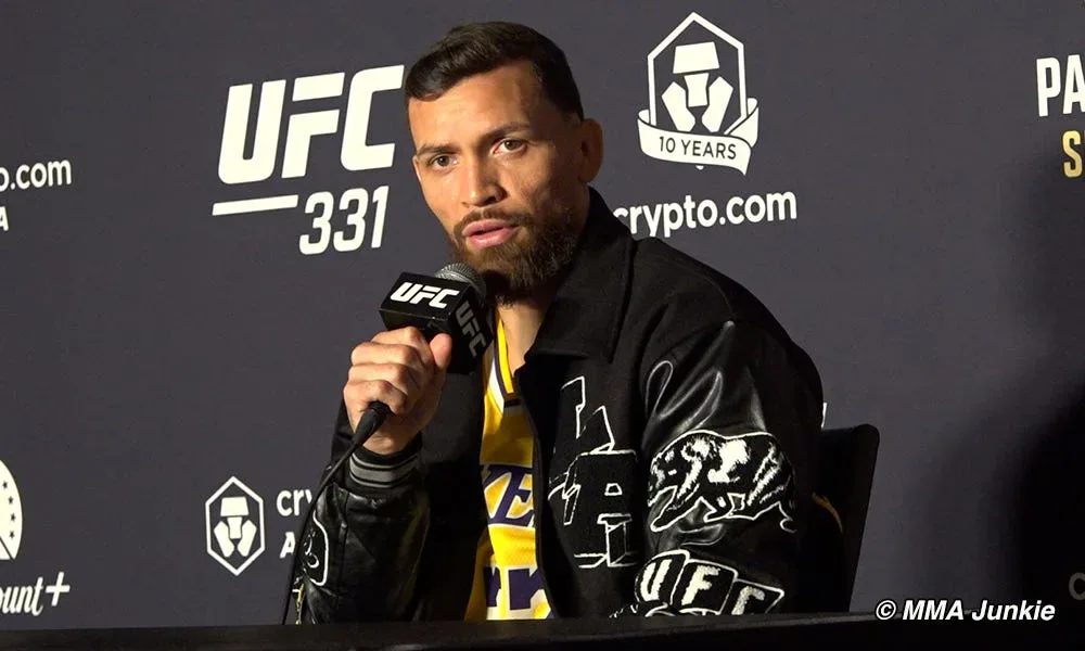 Lutador comparece a coletiva de imprensa do UFC 331 segurando microfone em frente a painel com logos do evento