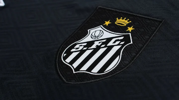Detalhe do escudo e da gola de camisa preta com listras brancas verticais, exibindo coroa e estrelas em dourado na parte superior do brasão