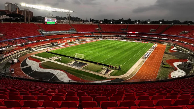 Estádio com arquibancadas vermelhas e gramado bem cuidado é visto vazio sob céu nublado