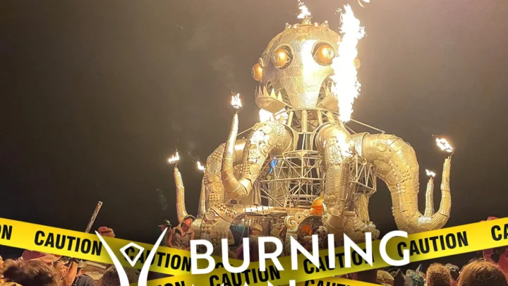 Estrutura de metal dourado com chamas saindo da cabeça e do corpo durante apresentação noturna no festival Burning Man, cercada por fitas de advertência amarelas