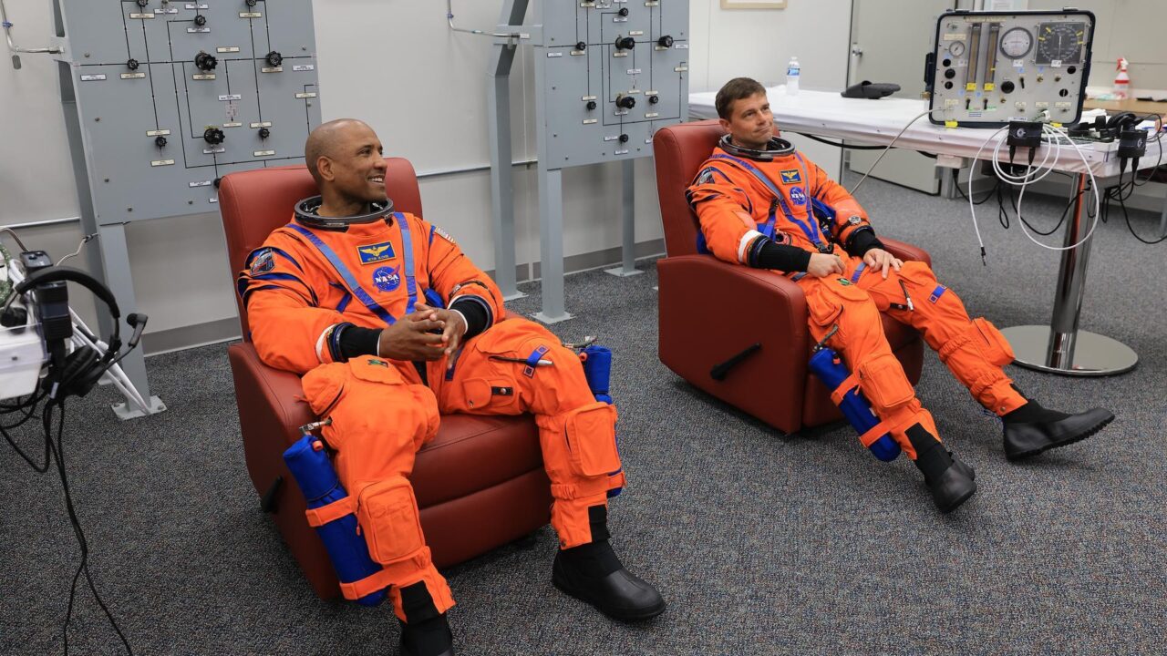 Da esquerda para a direita, os astronautas da NASA Victor Glover, piloto da Artemis II, e Reid Wiseman, comandante da Artemis II, realizam verificações de vazamento em seu traje espacial dentro da sala de vestimenta da equipe no Neil A. Armstrong Operations and Checkout Building, no Kennedy Space
