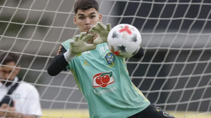 Goleiro da seleção brasileira realiza defesa durante treinamento ou partida, posicionado na frente da meta com as mãos estendidas para aparar a bola