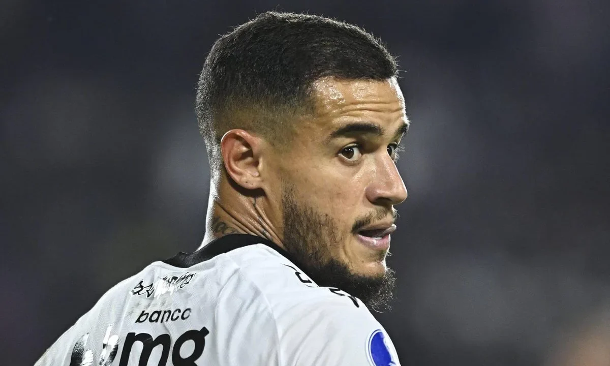 Jogador de futebol com uniforme branco de clube brasileiro com patrocínio de banco durante partida noturna