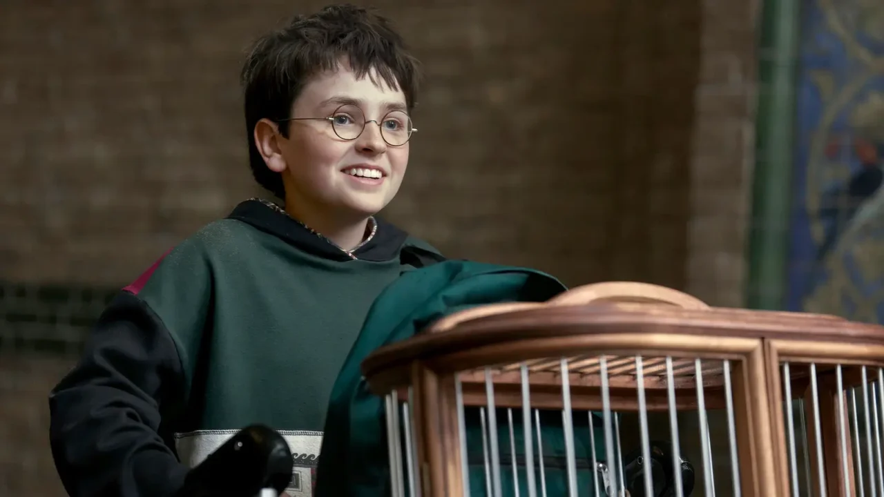 Menino de óculos em uniforme de escola de magia sorri enquanto segura uma gaiola de madeira em cena de produção audiovisual