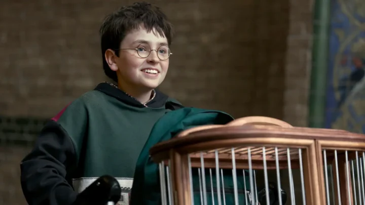 Menino de óculos em uniforme de escola de magia sorri enquanto segura uma gaiola de madeira em cena de produção audiovisual