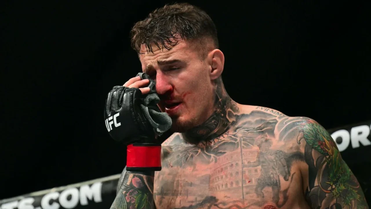 Lutador de MMA com luvas de competição UFC e tatuagens no tórax fala durante evento do UFC, com o rosto tocando a mão próximo à boca