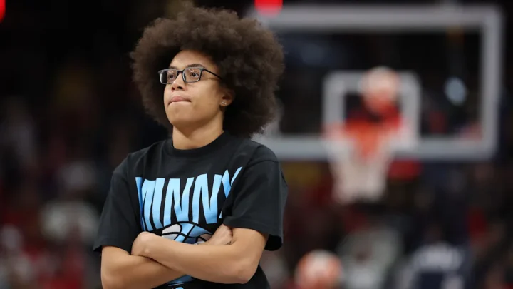 Jovem de óculos com camiseta preta estampa azul cruza os braços enquanto assiste a jogo de basquete em quadra