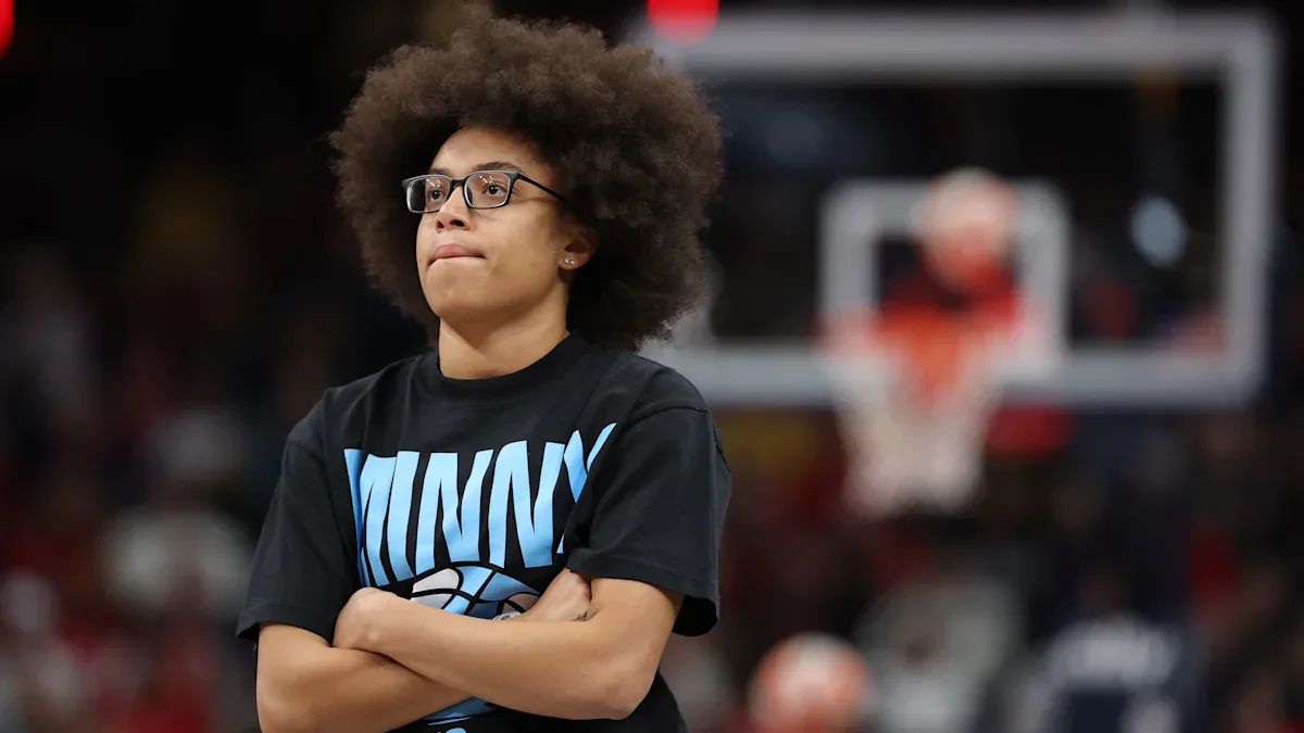 Jovem de óculos com camiseta preta estampa azul cruza os braços enquanto assiste a jogo de basquete em quadra