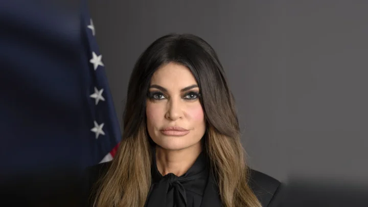 Retrato oficial de Kimberly Guilfoyle como embaixadora dos EUA na Grécia, de blusa preta, diante de uma bandeira americana