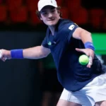 Tenista de camiseta azul-marinho com detalhe da bandeira do Brasil devolve bola de backhand com raquete vermelha em quadra de piso verde