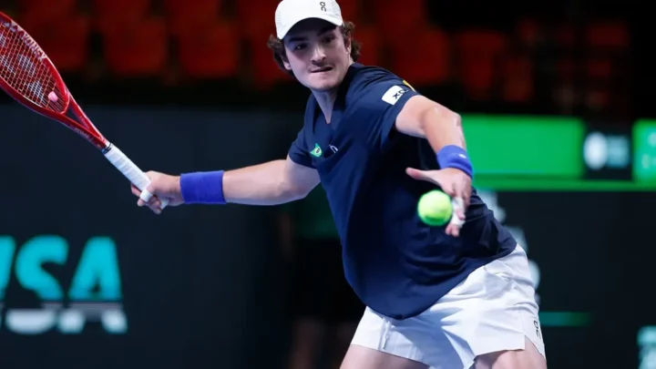 Tenista de camiseta azul-marinho com detalhe da bandeira do Brasil devolve bola de backhand com raquete vermelha em quadra de piso verde