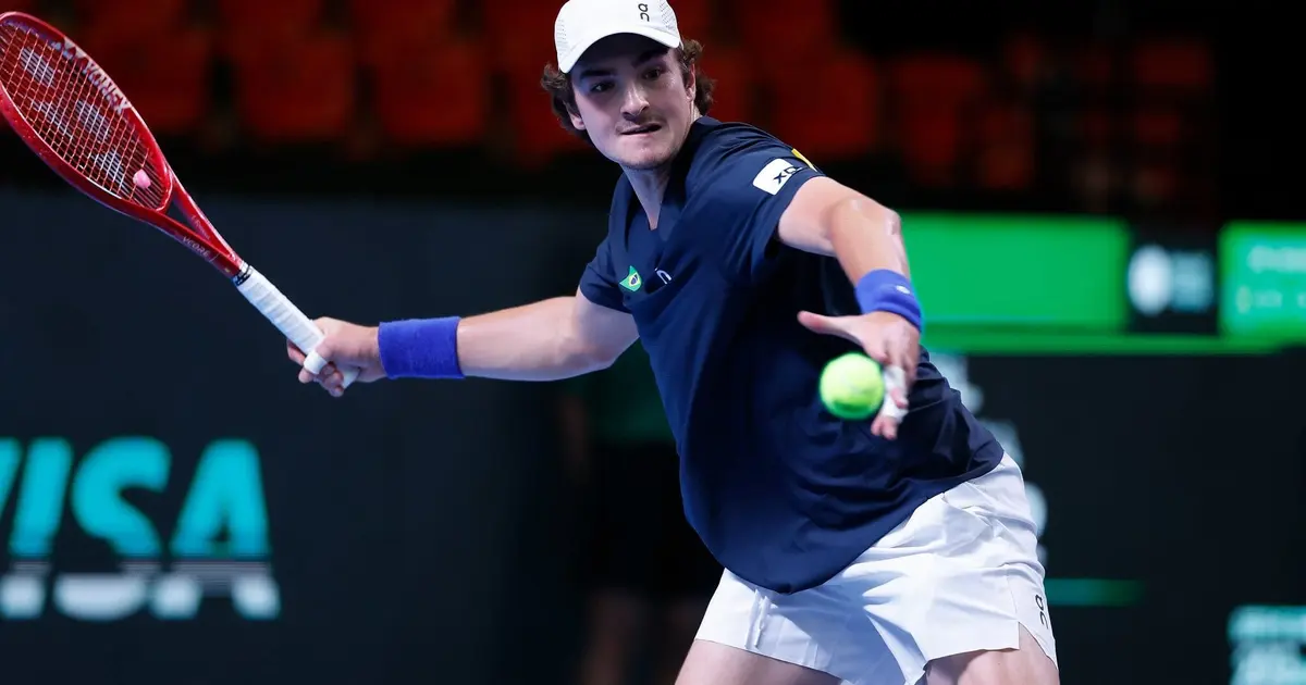 Tenista de camiseta azul-marinho com detalhe da bandeira do Brasil devolve bola de backhand com raquete vermelha em quadra de piso verde