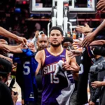 Jogador do Phoenix Suns, de camisa roxa e número 15, caminha por corredor ladeado por torcedores que estendem braços, camisas e canetas para autógrafos
