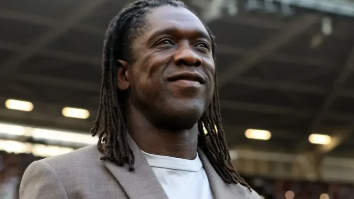 Homem de dreadlocks longos, com blazer bege sobre camiseta clara, sorrindo e olhando para o alto em um estádio com arquibancadas ao fundo