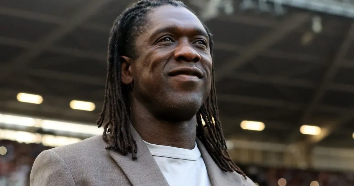 Homem de dreadlocks longos, com blazer bege sobre camiseta clara, sorrindo e olhando para o alto em um estádio com arquibancadas ao fundo