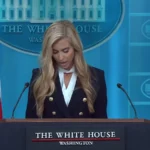 Katie Zacharia lê o discurso de abertura no púlpito da sala de imprensa da Casa Branca, ao lado da bandeira dos EUA, em 13 de outubro de 2026