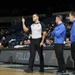Árbitro de camisa listrada levanta o dedo indicador enquanto jogador de camisa azul e branca, número 55, observa a marcação em quadra coberta