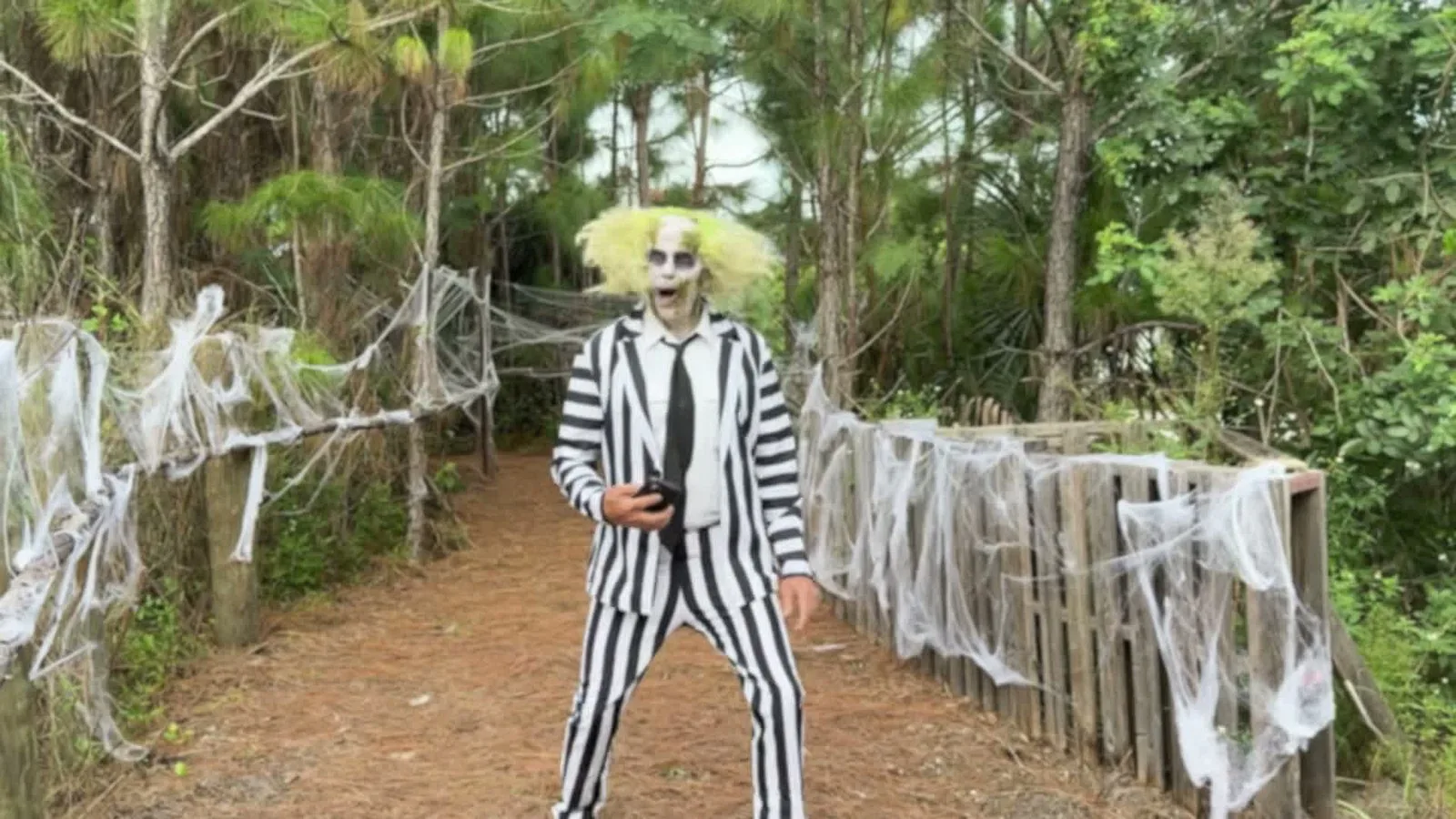 Beetlejuice, com o celular na mão, numa trilha decorada com teias de aranha