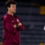 Homem de jaqueta vinho com logo de clube observa jogo em campo, com os braços cruzados