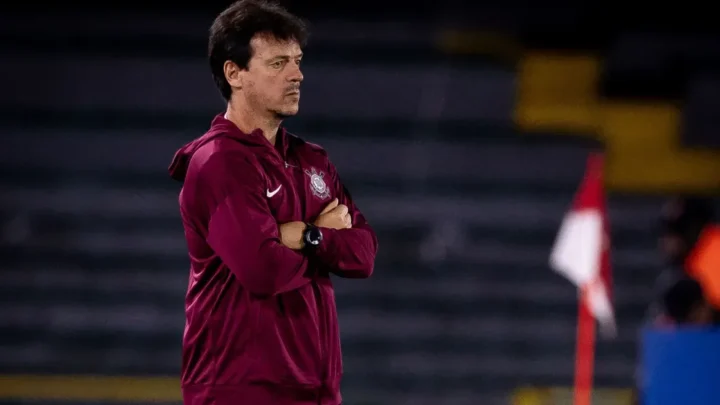 Homem de jaqueta vinho com logo de clube observa jogo em campo, com os braços cruzados