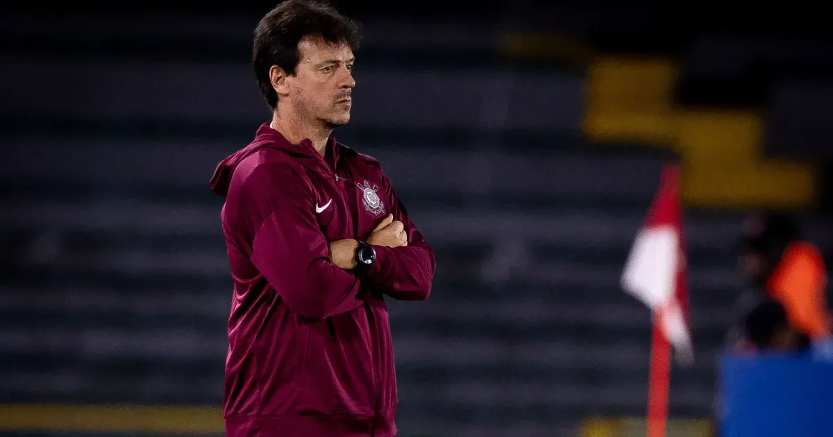 Homem de jaqueta vinho com logo de clube observa jogo em campo, com os braços cruzados