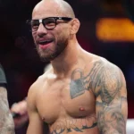 Lutador sem camisa e de óculos escuros sorri ao lado de um ringue do UFC, com tatuagens no peito e nos braços