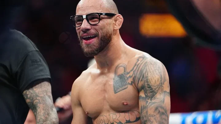 Lutador sem camisa e de óculos escuros sorri ao lado de um ringue do UFC, com tatuagens no peito e nos braços