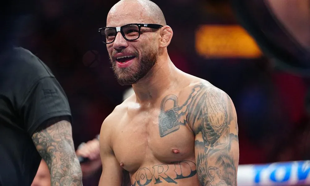 Lutador sem camisa e de óculos escuros sorri ao lado de um ringue do UFC, com tatuagens no peito e nos braços