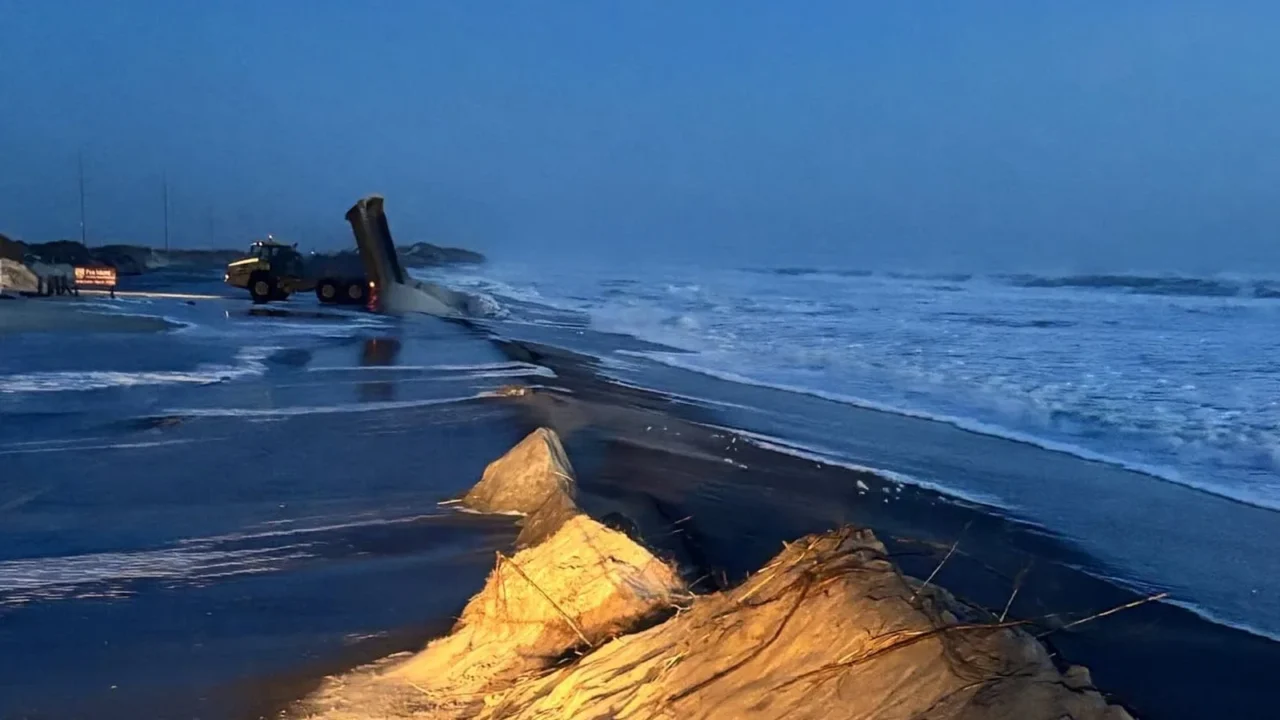 Duna de areia desfeita pelo mar ao lado da rodovia NC 12 molhada, com o oceano agitado e uma máquina de limpeza ao fundo, em Pea Island