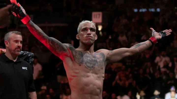 Lutador de MMA com cabelo loiro e tatuagens no peito e braços ergue os braços em arena lotada, com um integrante da equipe do UFC ao lado
