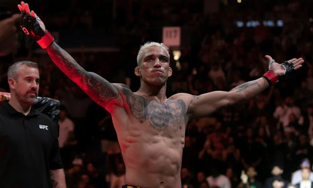 Lutador de MMA com cabelo loiro e tatuagens no peito e braços ergue os braços em arena lotada, com um integrante da equipe do UFC ao lado