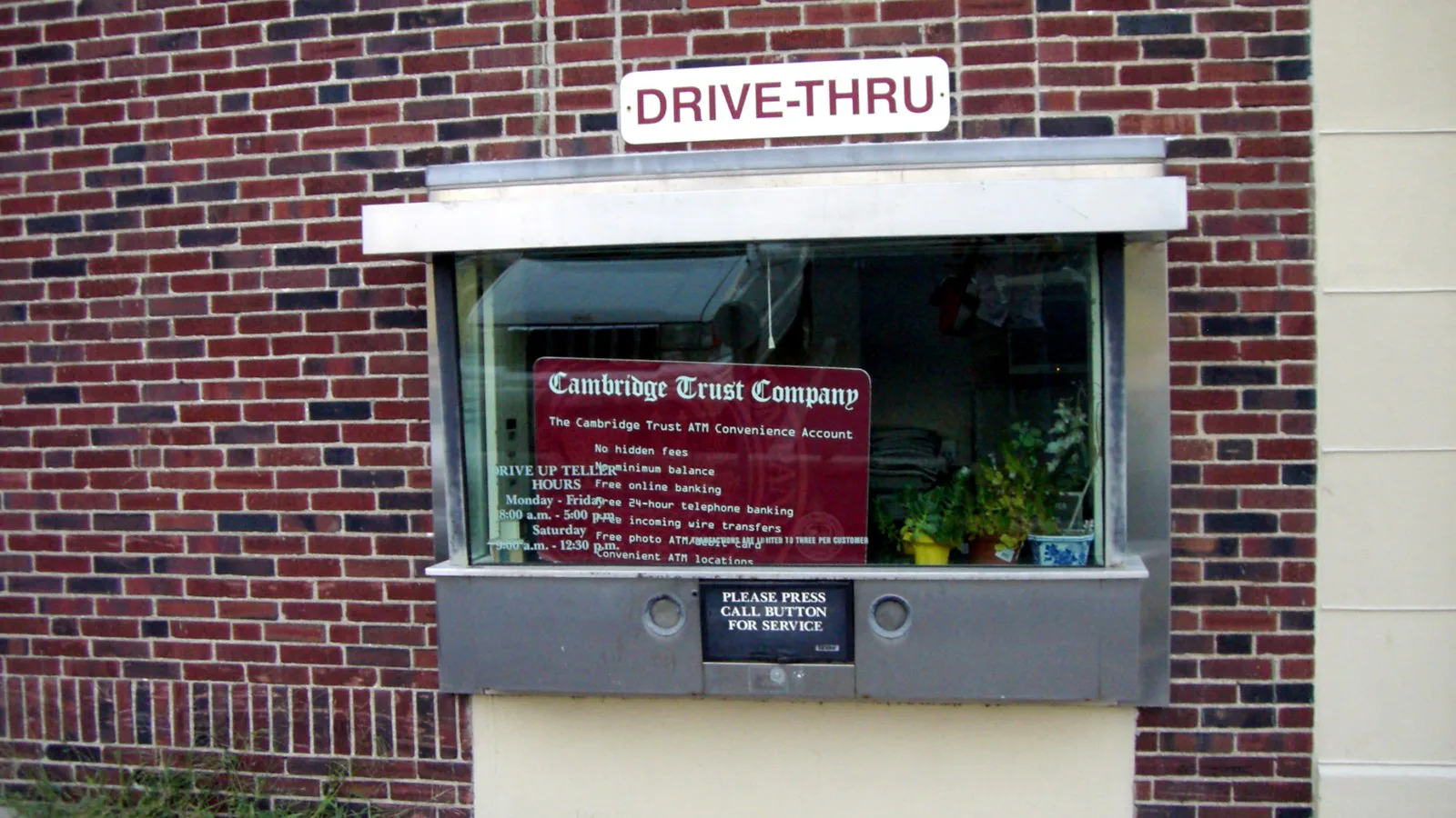 Janela de atendimento drive-thru numa parede de tijolos, com placa "Drive-Thru" e avisos do banco no vidro (imagem ilustrativa de 2006, sem relação com o vídeo)