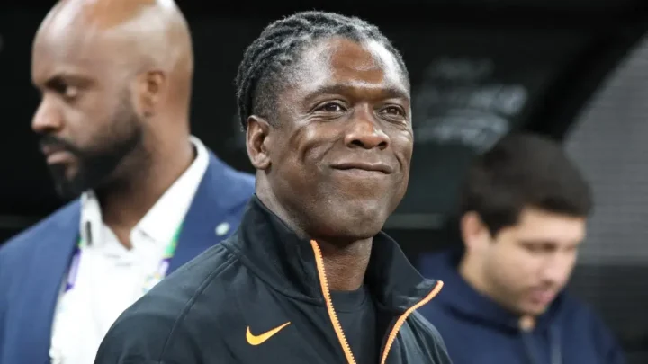 Homem de jaqueta preta com zíper laranja e logo da Nike sorri em um ambiente de estádio, com outras pessoas ao fundo