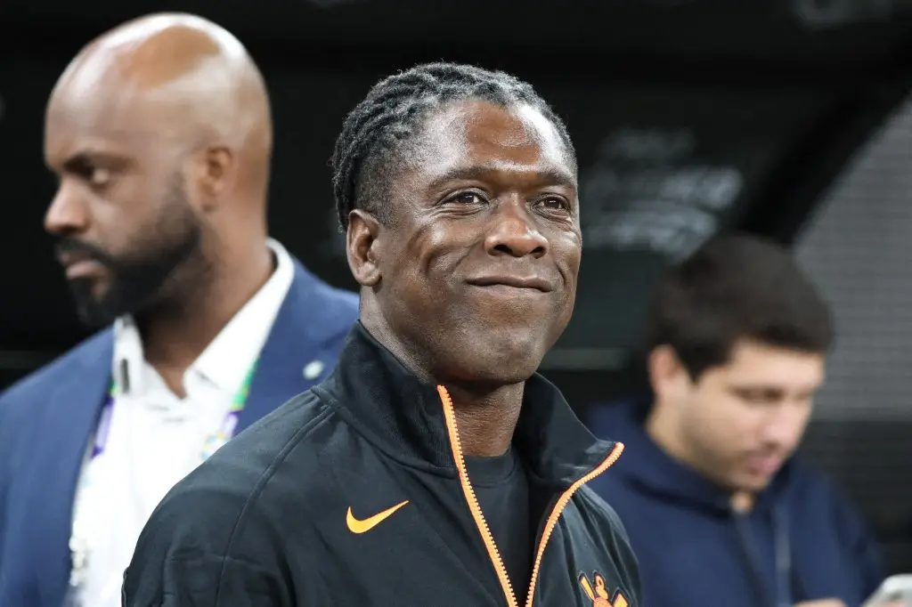Homem de jaqueta preta com zíper laranja e logo da Nike sorri em um ambiente de estádio, com outras pessoas ao fundo