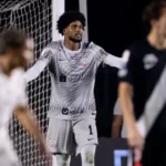 Jogador do Corinthians, de uniforme cinza com o número 1, gesticula em campo ao lado de uma rede de gol, enquanto atletas de camisa preta e branca aparecem desfocados em primeiro plano